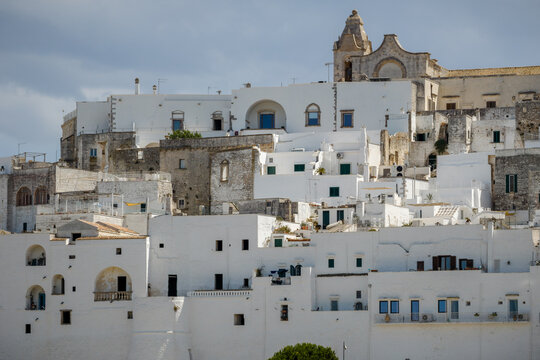 Aerial view of a cascade of dazzling white buildings, crowned by the Ostuni Cathedral against a backdrop of soft blue skies, Ostuni, Apulia, Italy.
