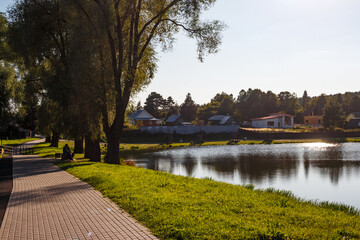 A peaceful lakeside promenade with a person relaxing on a bench under lush trees. Sunlight reflects off the serene water, creating a tranquil outdoor scene perfect for a leisurely stroll