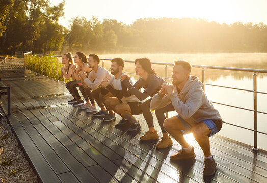 Positive people perform squats during group training in city park outdoors, boosting sport teamwork, engaging in fitness exercise, uniting team in a shared workout experience on urban green grounds.