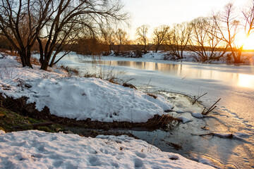 Serene winter river landscape with snow-covered banks, partially frozen water, and a small spring. Golden hour light bathes the scene, highlighting bare trees and tranquil reflections
