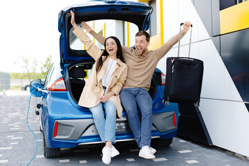 Young couple man and woman traveling by electric car having stop at charging station. Boyfriend and girlfriend sitting in the car trunk and smiling