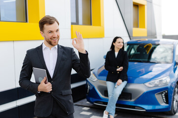 Young handsome car dealership consultant holding a tablet while standing near the car and showing hand gesture ok
