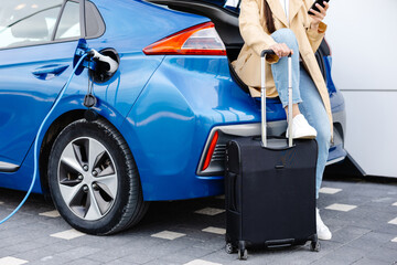 Young woman waiting charging automobile battery from small public station and using smartphone while charging automobile