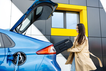 A young woman stands next to an electric car while charging the car at the station and puts her luggage in the trunk