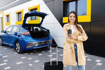 Young woman with cup of coffee waiting while her electric car charging, sustainable and economic transportation concept