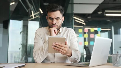 Thoughtful businessman is using digital tablet thinking about solving a problem sitting at workplace in business office. Serious focused manager busy with project or working on a challenge. Front view