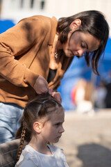 Vertical photo of a mother making a braided hairstyle for her daughter while sitting together at an outdoor table, concept of care and family tenderness.