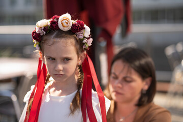 Sad girl with braided hair and a floral wreath with ribbons, with her mother in the background helping her get dressed, concept of care and emotional connection.