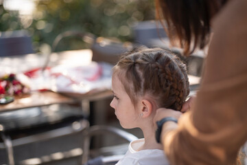 Horizontal rear view of a mother making a braided hairstyle for her daughter while sitting together at an outdoor table, concept of care and family tenderness.