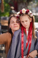 Vertical photo of a dark-haired mother in a brown sweater hugging her sad daughter in a large gray jacket with a floral wreath and ribbons, concept of love, care, and emotional support.