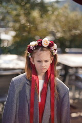 Vertical photo of a lonely sad girl in a large gray jacket with a floral wreath and ribbons looking down outdoors, concept of childhood and solitude.