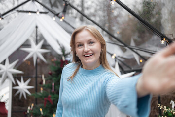 Smiling blonde Caucasian woman in a light blue sweater taking a selfie in cozy festive Christmas interior. Holiday decorations, Christmas tree, and glowing lights create a joyful and cheerful winter