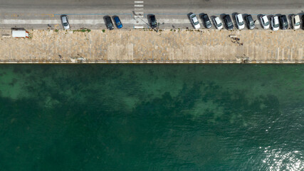 Perpendicular aerial view of a seafront. There is a sidewalk, a road, and parked cars.