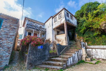 A charming, weathered stone building with a wooden balcony and vibrant pink flowers stands on a cobblestone street