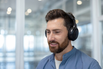Bearded man smiling and concentrating, wearing headphones and enjoying audio content in a modern office environment, representing focus and productivity
