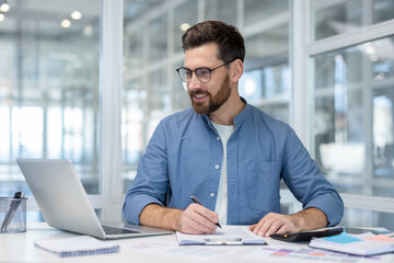 Young man with glasses smiling while taking notes and using a laptop at his desk in a modern office, focused and confident while reviewing documents and planning work