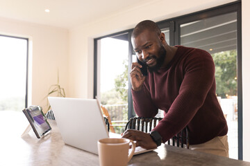 African American man leaning on kitchen countertop holding phone and typing on laptop, copy space
