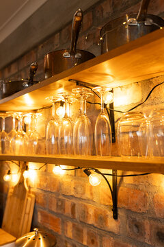 Wooden shelves holding metal pans with hanging wine glasses on brick wall, string lights glowing