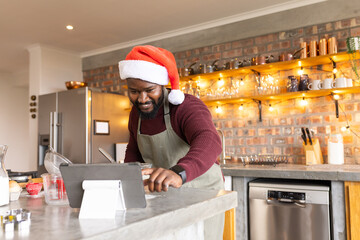 African American man pointing at tablet on kitchen island while wearing Santa hat and apron