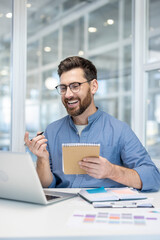 Smiling man engaging in a video conference from his modern office desk, holding a notebook and pen while actively participating in an online business meeting or distant learning session