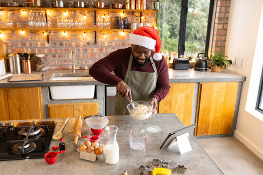 African American man mixing dough at kitchen island wearing Santa hat and apron with digital tablet