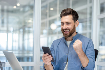 Happy businessman pumps his fist in celebration while reading excellent news on his smartphone in a...
