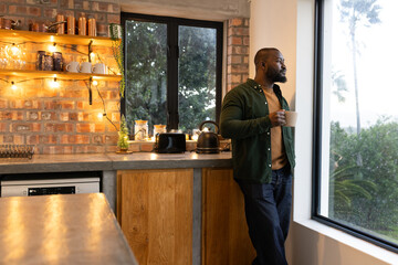 African American man holding coffee mug, gazing out window by kettle on kitchen counter, copy space