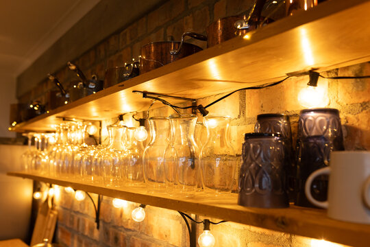 Wooden shelves are holding glass carafes, mugs and copper pots under filament lights on brick wall