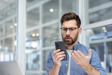 Man in an office setting looking confused and frustrated while holding a smartphone, reacting to bad news or a complex problem causing stress and disappointment