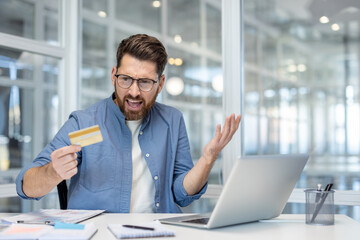 Frustrated man experiencing banking problems, holding a credit card and using a laptop in an office, facing issues with an online payment or a failed transaction