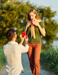 Obraz premium Cheerful handsome man proposing to his girlfriend. Portrait of a young happy couple having fun outdoors in nature during sunset. Girlfriend and boyfriend bonding, love concept during summer.