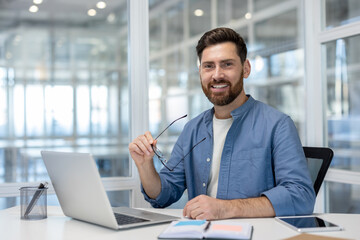Smiling bearded man in a casual blue shirt and white t-shirt sitting at a contemporary office desk, holding his eyeglasses, and looking directly at the camera with a laptop and tablet nearby