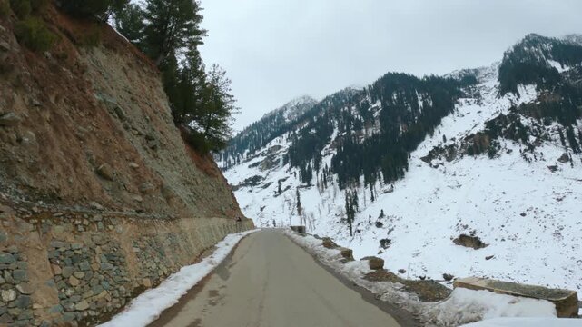 4K POV shot of a vehicle driving through a narrow asphalt road on the side of the Himalayan mountains covered by snow in winter season towards Chandanwari from Pahalgam in Jammu and Kashmir, India.