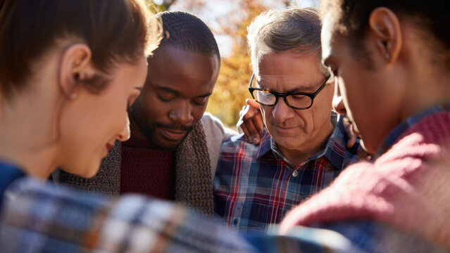 Diverse group of people standing in a circle and praying outdoors during a sunny autumn day, heads bowed, eyes closed, expressing unity, spirituality, and support through a moment of faith