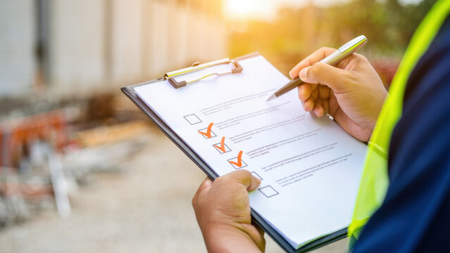 Construction checklist on a clipboard, safety inspection on site, worker in high visibility vest, quality control, compliance review, project management, task verification, pen writing, file