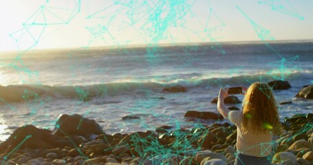 Capturing woman holding smartphone on pebble beach at sunset, turquoise network overlay, copy space