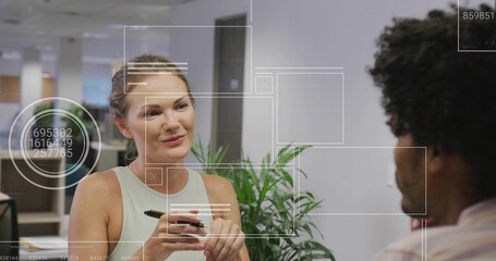 Explaining female office worker using pen outlining point to colleague in open-plan office