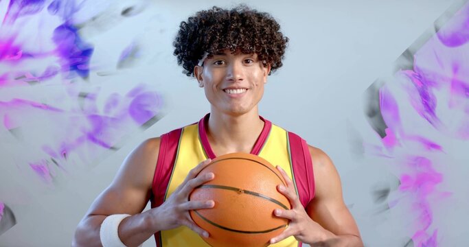 Athlete wearing jersey holding orange basketball smiling against studio backdrop with purple shapes - Powered by Adobe