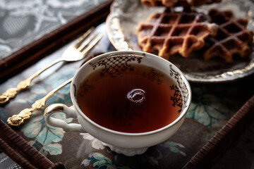 Elegant cup of tea on a floral tray with golden spoons and waffles on a vintage plate, creating a...