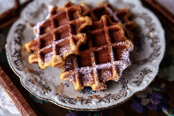Close-up of golden crispy waffles sprinkled with powdered sugar on an elegant vintage plate, creating a cozy homemade breakfast atmosphere.