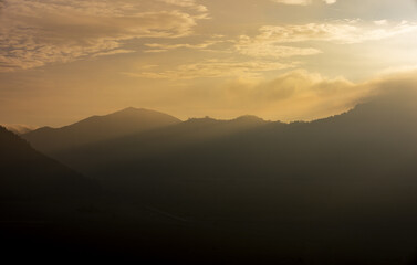 View of a golden sunrise casts an ethereal glow over the silhouetted mountains, shrouded in a mysterious mist, creating a serene landscape, Bromo, East Java, Indonesia.