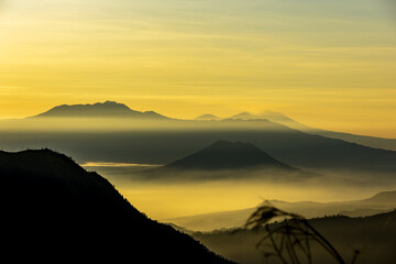 View of golden light filtering through the misty valleys and silhouetted mountains, creating a layered landscape of warmth and shadow, Bromo, East Java, Indonesia.