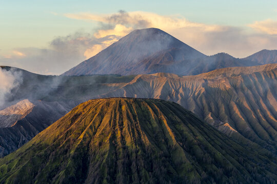 View of the imposing volcanic landscape, with its rugged peaks and shadowed valleys, shrouded in ethereal mist under a tranquil sky, Bromo, East Java, Indonesia.