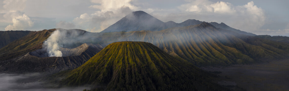 View of rugged volcanic peaks, shrouded in mist and kissed by golden light, rise majestically in a panoramic vista, Bromo, East Java, Indonesia.