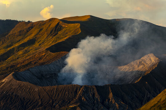 View of billowing white smoke rising above the dark, textured crater of a volcano, set against the warm glow of a mountain range, Bromo, East Java, Indonesia.