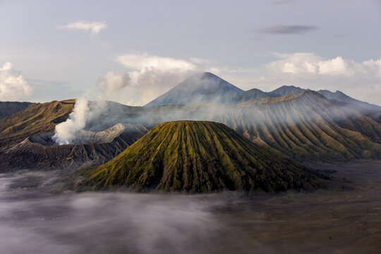 View of a majestic volcano shrouded in ethereal mist, its rugged terrain contrasting with the soft clouds, a scene of raw natural beauty, Bromo, East Java, Indonesia.
