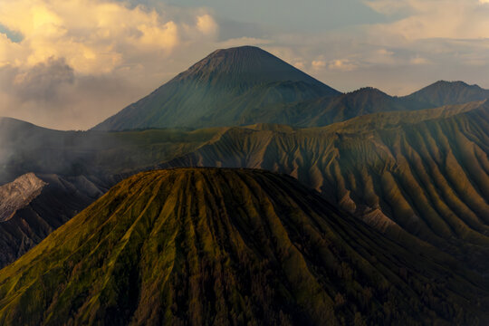 View of majestic, tiered volcanic peaks rise against a backdrop of soft, golden-tinged clouds, casting long shadows across the rugged landscape, Bromo, East Java, Indonesia.