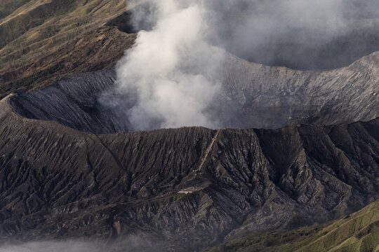 View of the rugged, dark volcano crater exhales a plume of white smoke against the textured slopes, a stark contrast, Bromo, East Java, Indonesia.