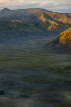 View of the misty valley floor stretches toward rugged mountains, illuminated by the sun's golden touch, contrasting with the cool shadows, Bromo, East Java, Indonesia.