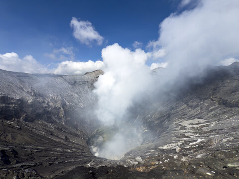 View of volcanic crater spewing white smoke under a sky dotted with fluffy clouds, showcasing the stark contrast of textures, Bromo, East Java, Indonesia.